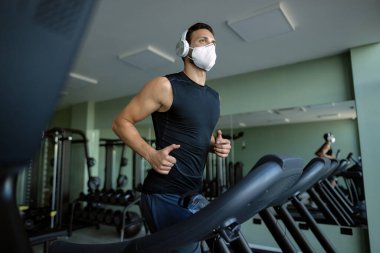 Low angle view of athletic man wearing protective face mask while running on treadmill in a gym. 