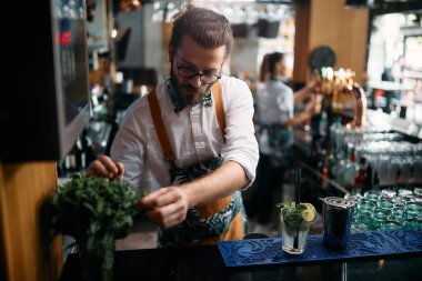 Young bartender using mint herb while preparing mojito cocktail at bar counter.