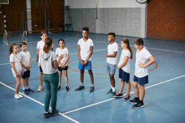 Large group of elementary students listening to their PE teacher during physical activity class at school gym. 