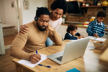 African American man writing notes and working on laptop while being at home with his wife and kids. 