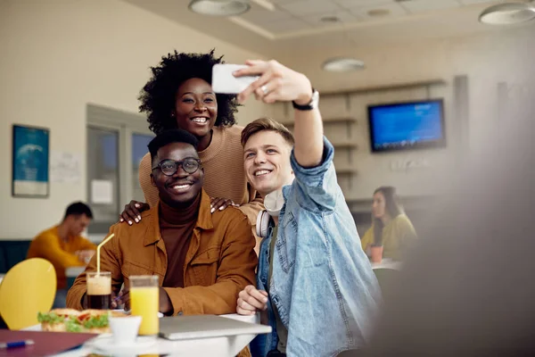 Happy university friends having fun while taking selfie with smart phone in a cafeteria.