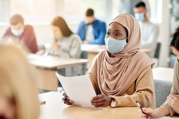 Group of college students having a class at lecture hall during COVID-19 pandemic. Focus is on Muslim female student with protective face mask.