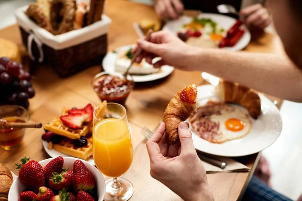 Close-up of man having croissant with jam while eating breakfast with his girlfriend at dining table.