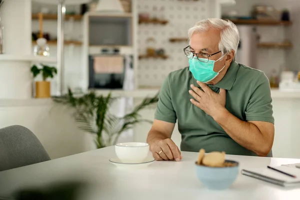 Senior man with a face mask coughing and holding his chest in pain at home.