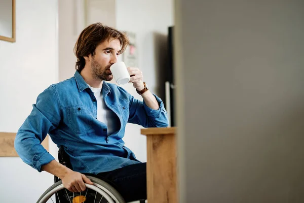pensive disabled man drinking tea while being in wheelchair at home. Copy space.