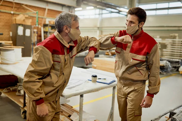 Happy carpenters elbow bumping while working in a workshop during coronavirus pandemic.