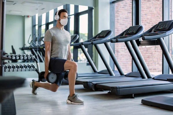 Male athlete exercising with hand weights in lunge position while wearing protective face mask in health club.