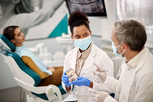 African American dentist using dentures and talking to her colleague while having appointment with female patient at dental clinic.