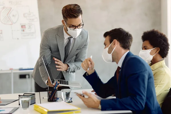 Young entrepreneur using laptop while talking to his coworkers during business meeting in the office. They are wearing protective face masks due to coronavirus epidemic.