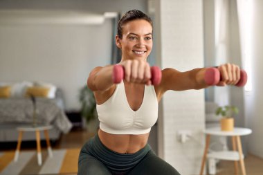 Happy sportswoman doing squats while exercising with hand weights at home. 
