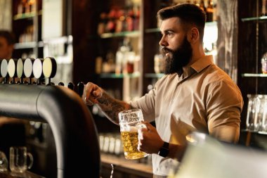 Young bartender pouring beer from beer tap while working in a pub. 