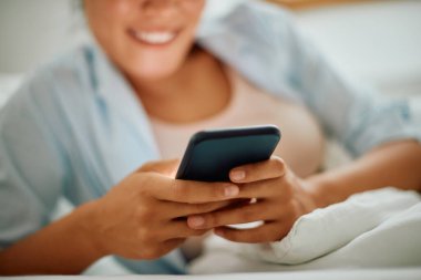 Close-up of woman typing text message on mobile phone while relaxing on a bed. 