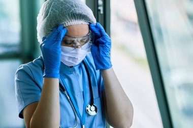 Overworked healthcare worker holding her head in pain at the hospital during COVID-19 epidemic.
