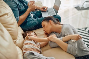 Young mother spending time with her son while father is using laptop at home.