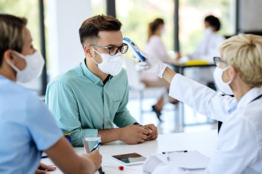 Young man wearing protective face mask while doctor is measuring his temperature with infrared thermometer during appointment at medical clinic. 