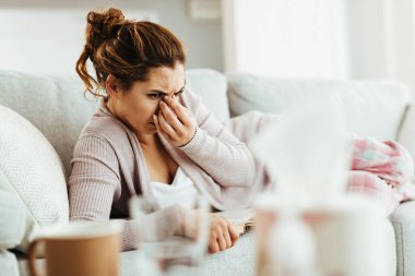 Young woman holding her head in pain while lying down on sofa and reading book at home. 