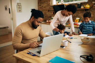 Black working father using laptop and analyzing paperwork while mother is assisting their kid with learning at home.