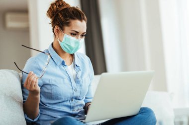 Smiling woman wearing face mask while surfing the net on laptop and relaxing at home during virus epidemic. 