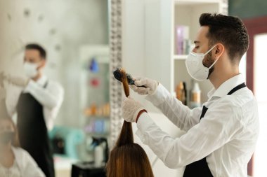 Young hair stylist wearing protective face mask while brushing woman's hair and working during coronavirus epidemic.