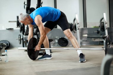 Mature athlete adding weight disk on barbell while preparing for weightlifting in a gym. 