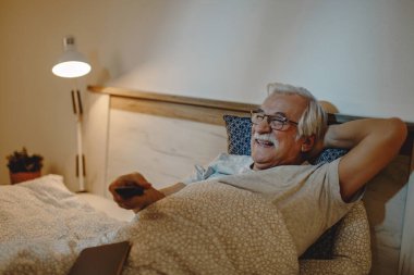 Happy senior man watching TV while relaxing in bedroom in the evening. 