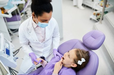 Small girl holding dentist's hand and complaining about toothache during appointment at dental clinic. 