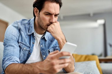 Young man feeling worried while reading text message on mobile phone at home.