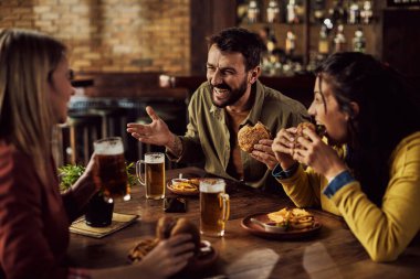 Group of happy friends having fun and talking while eating hamburgers and drinking beer in a pub. Focus is on man.