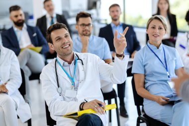 Happy doctor attending a seminar and raising his hand to ask a question at convention center. 