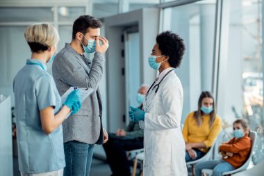African American doctor talking to a patient who is complaining of headache while wearing face masks and standing in a hallway at the clinic.  