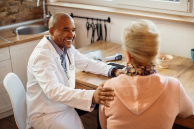 Happy African American general practitioner communicating with senior woman during home visit. 