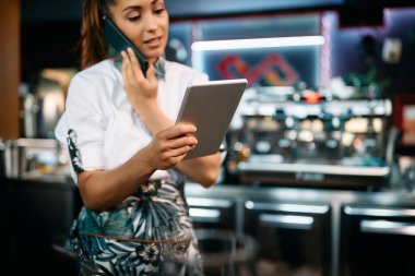 Close-up of waitress using digital tablet while communicating on cell phone in a pub. 
