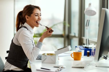 Young happy businesswoman eating while using desktop PC in the office. 