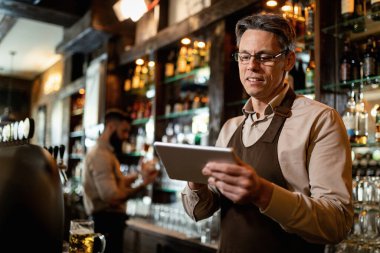 Smiling barista working on digital tablet in a pub. His coworker is in the background. 
