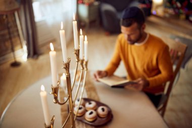 Close-up of lighted candles in menorah with Jewish man reading Hebrew bible at home.