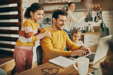 Happy man working at home on a computer while his daughters are around him. Mother is in the background. 