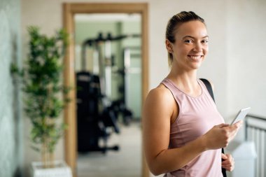 Young happy sportswoman using mobile phone in a lobby at the gym and looking at camera. 