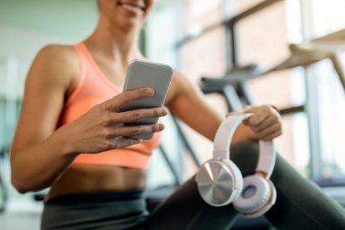 Close-up of athletic woman texting on smart phone while holding headphones and relaxing at the gym.