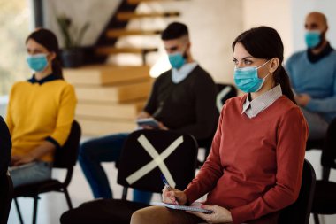 Female entrepreneur writing notes while attending business seminar and wearing face mask due to COVID-19 pandemic. 