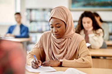 African American female student in hijab taking notes while attending a class at lecture hall.