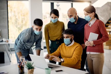 Group of colleagues cooperating while working on laptop and wearing protective face masks in the office. 