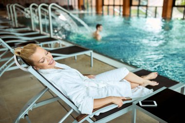 Smiling woman in bathrobe relaxing with eyes closed by the swimming pool at health spa. 