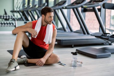 Pensive athlete taking a break from exercising while sitting on the floor in a gym. 