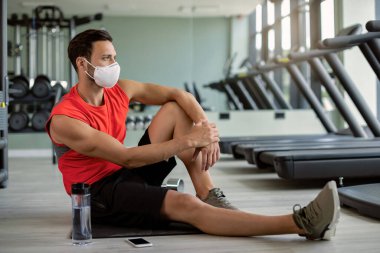 Athletic man wearing protective face mask while taking a break from exercising in a gym during coronavirus epidemic. 