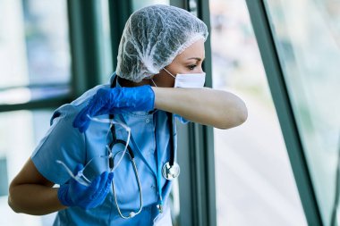 Nurse wearing protective workwear and sneezing into elbow while working at medical clinic during coronavirus epidemic. 