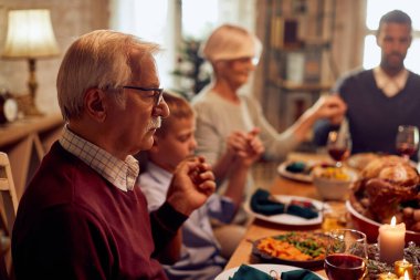 Multi-generation family praying on Thanksgiving while holding hands during a meal at dining table. Focus is on senior man.
