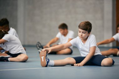 Schoolboy and his friends doing relaxation exercises and warming up for physical activity class at school gym. 