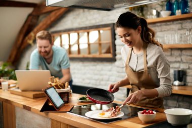 Happy woman making breakfast for her boyfriend who is using laptop in the background. 