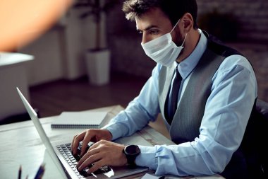 Businessman with face mask working on laptop in the office during coronavirus pandemic. 