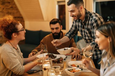 Young smiling man serving food while having dinner with his friends at dining table at home.